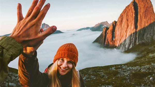 Насмеана жена во портокалова шапка ја крева раката кон лицето пред неа, со планини и море од облаци во позадина.