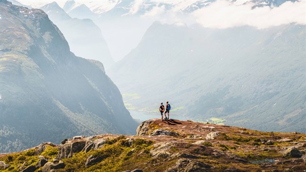 Двајца луѓе стојат на врвот на карпа и набљудуваат планински пејзаж покриен со магла.