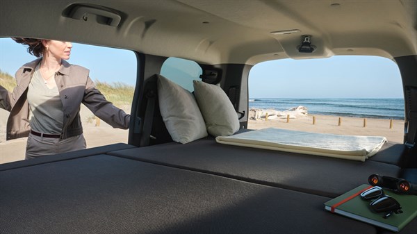 Внатрешноста на Dacia Jogger со поставен кревет и отворената задна врата со поглед на плажата.