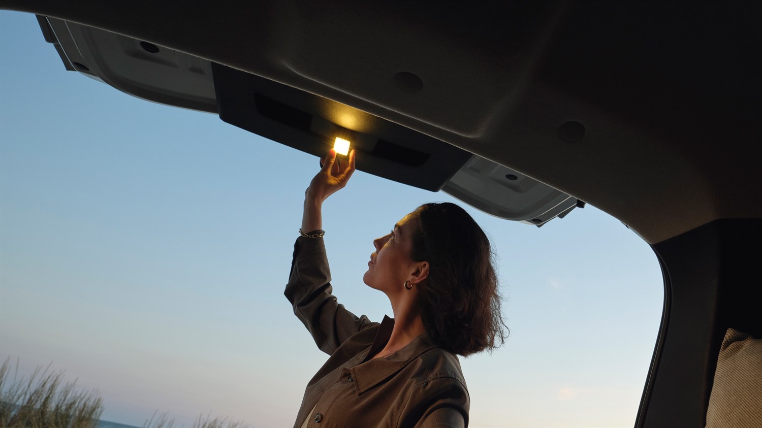 Жена во Dacia Jogger го вклучува внатрешното светло во отворениот багажен простор при зајдисонце.