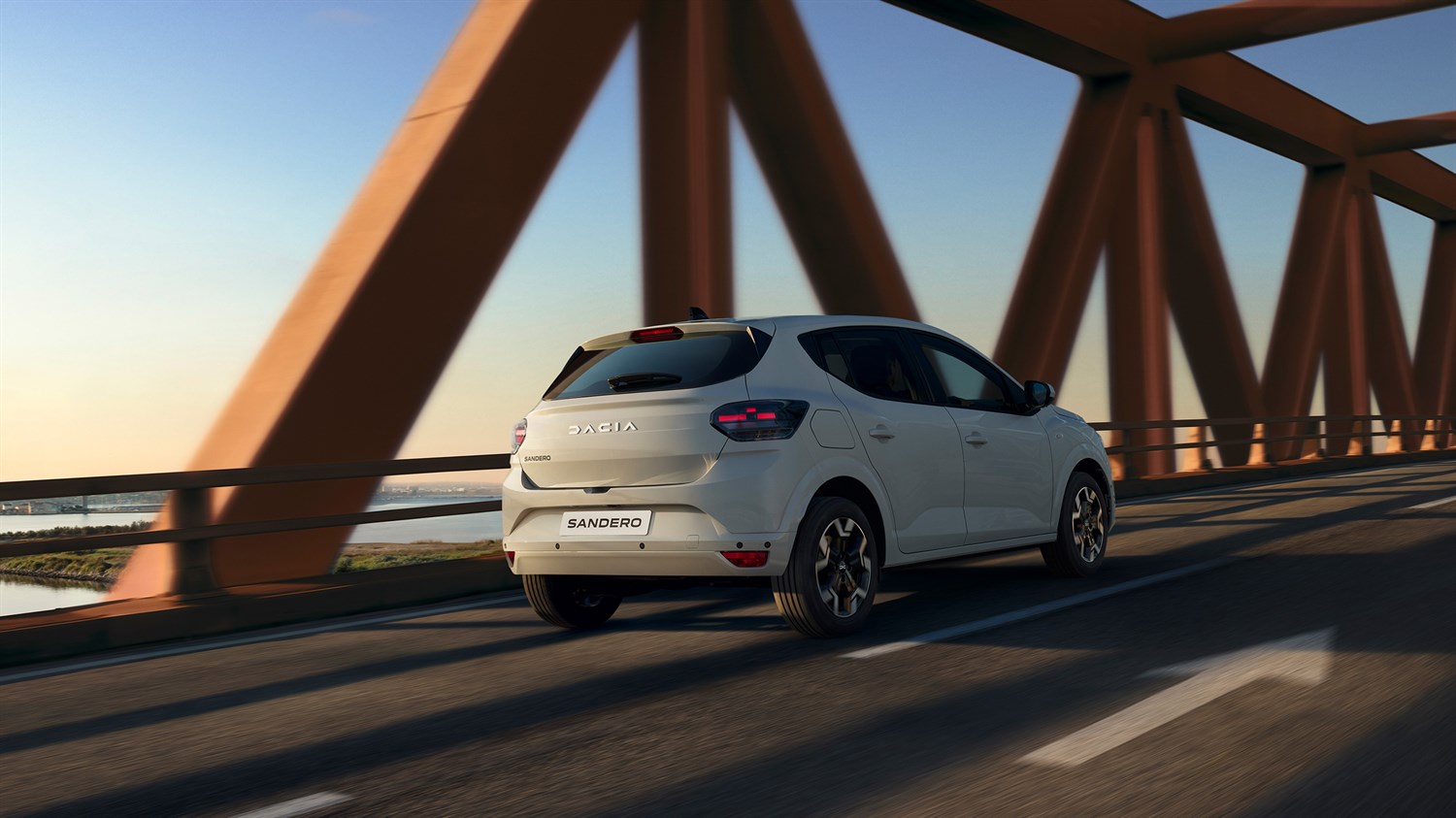 Dacia Sandero вози преку модерен мост при зајдисонце, прикажано одназад во движење.