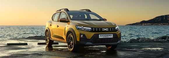 Dacia Sandero Stepway во златна боја паркиран покрај морето на зајдисонце, со планини во позадина.