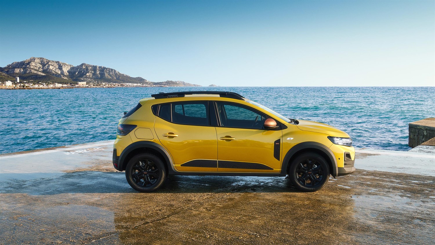 Vozilo Dacia Sandero Stepway u žutoj boji snimljeno iz profila uz more, parkirano na betonskoj površini s planinskim krajolikom u pozadini.