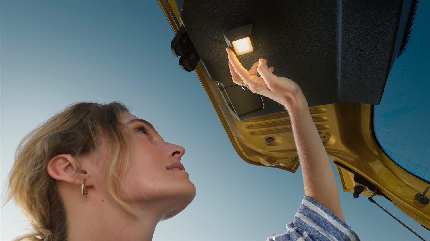 Жена ја крева раката кон внатрешното светло на отворениот багажен простор на Dacia Sandero Stepway, фотографирано од низок агол кон небото.